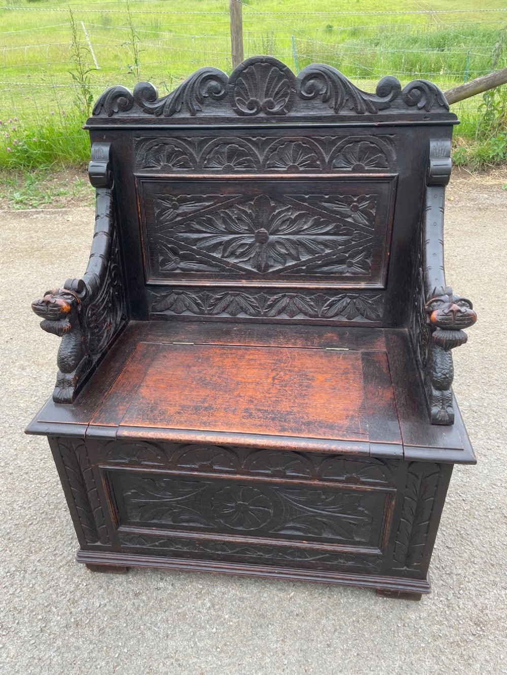 A 19th-century dark oak box settle with carved floral panels, lion mask terminals, a decorative cresting rail, and a lift-up storage seat. View XI