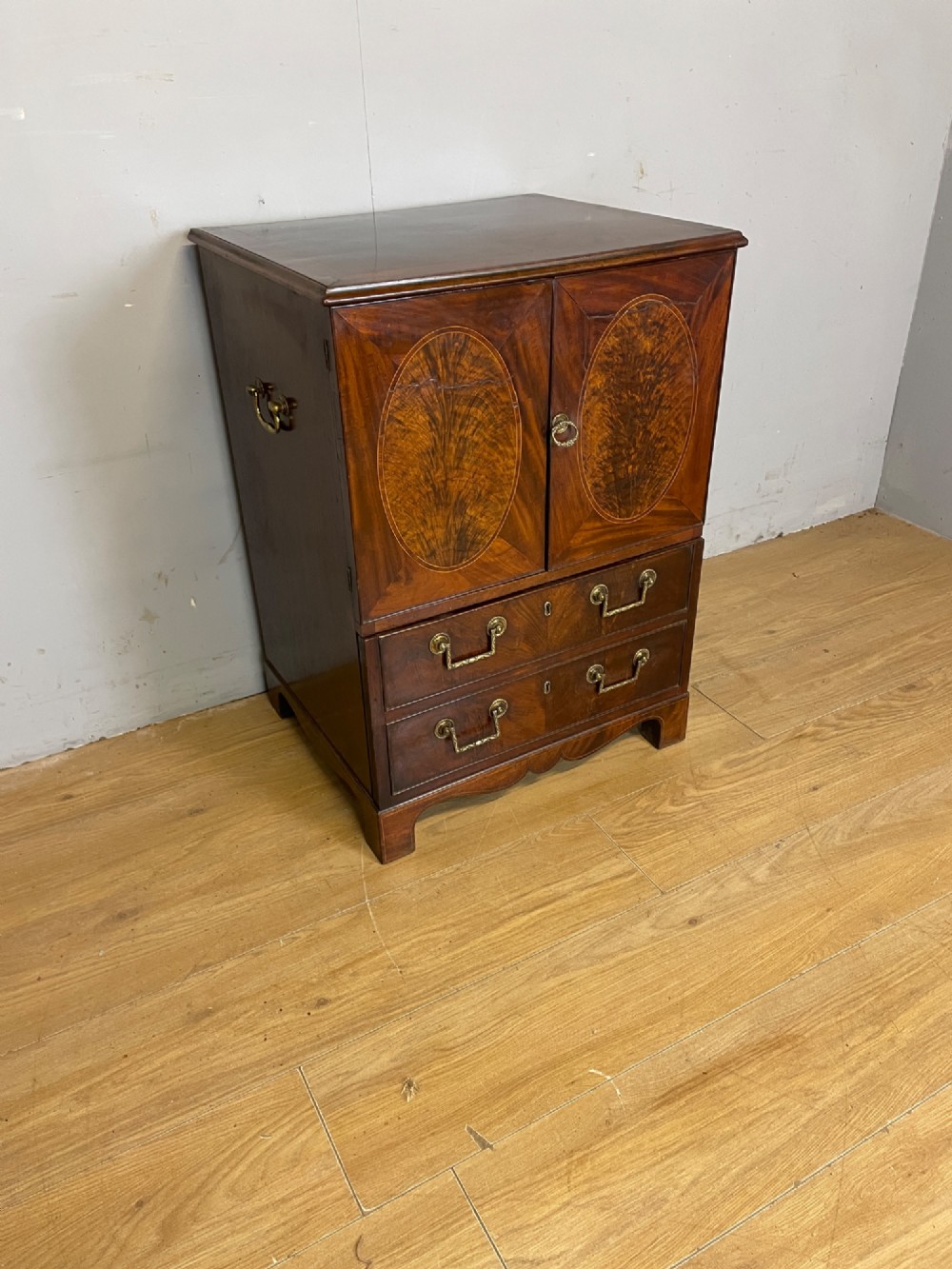 A Georgian mahogany side cabinet featuring oval flame veneer inlaid cupboard doors above two drawers with brass handles, resting on bracket feet.