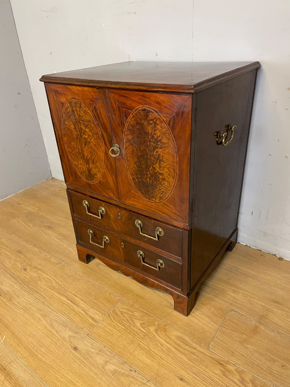 A Georgian mahogany side cabinet featuring oval flame veneer inlaid cupboard doors above two drawers with brass handles, resting on bracket feet. View III
