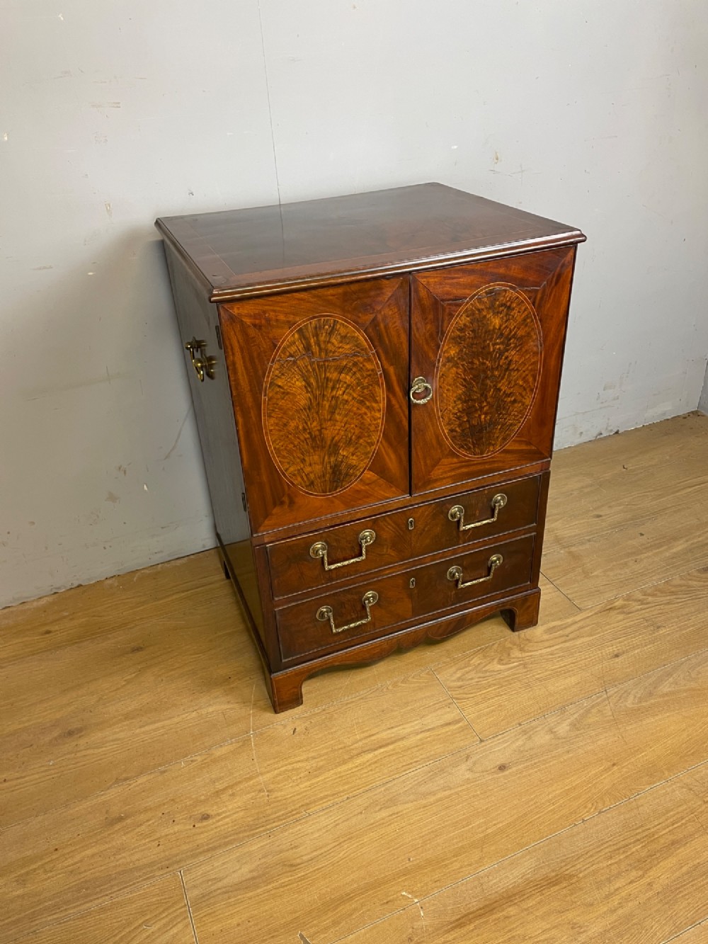A Georgian mahogany side cabinet featuring oval flame veneer inlaid cupboard doors above two drawers with brass handles, resting on bracket feet. View XI