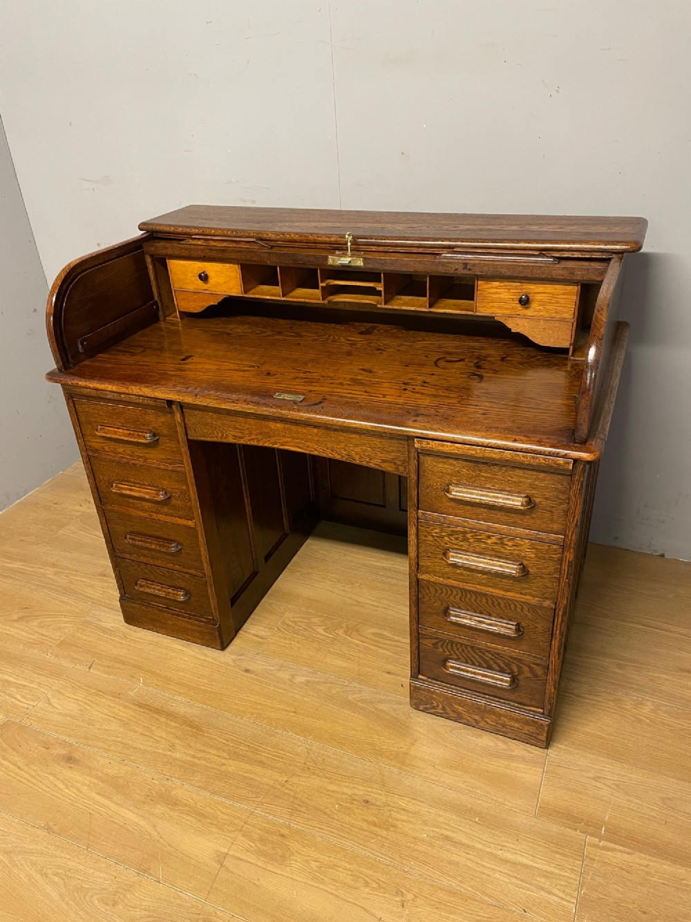 An Edwardian solid oak twin-pedestal roll top desk featuring a curved tambour front, fitted drawers with integrated wooden handles, and a central brass keyhole. View IV