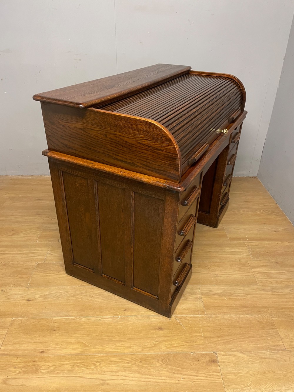 An Edwardian solid oak twin-pedestal roll top desk featuring a curved tambour front, fitted drawers with integrated wooden handles, and a central brass keyhole. View IX
