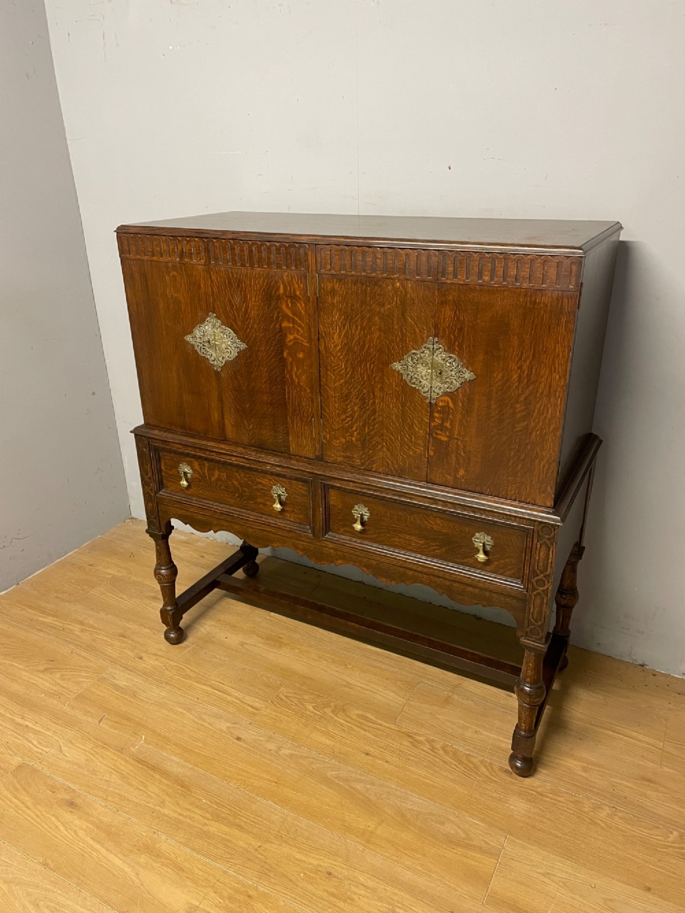 A solid dark oak Jacobean style side cabinet featuring brass escutcheons, carved moulding along the top edge, two lower drawers, and turned legs connected by a lower stretcher.