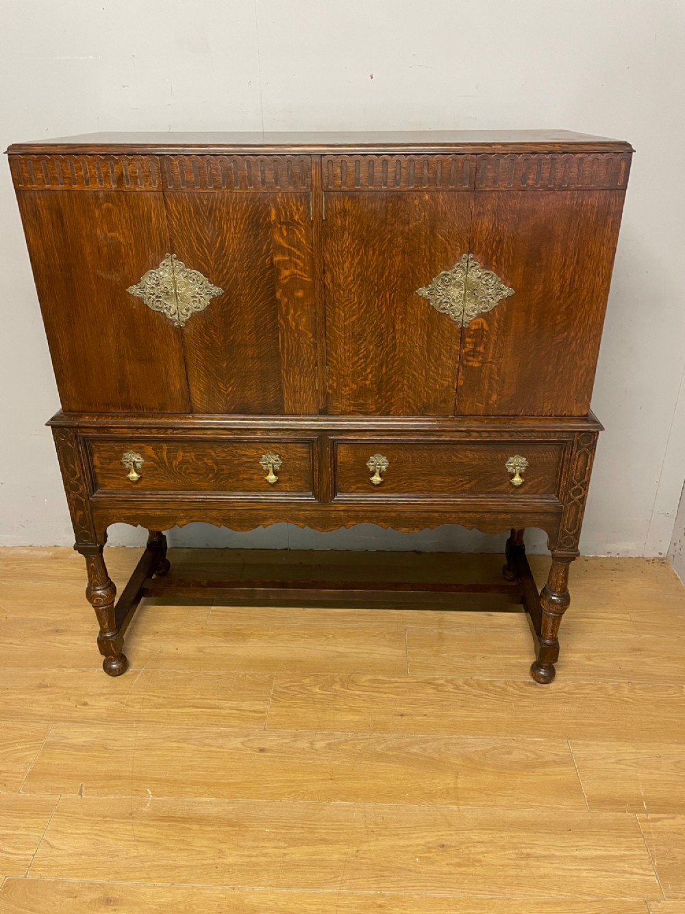 A solid dark oak Jacobean style side cabinet featuring brass escutcheons, carved moulding along the top edge, two lower drawers, and turned legs connected by a lower stretcher. View II