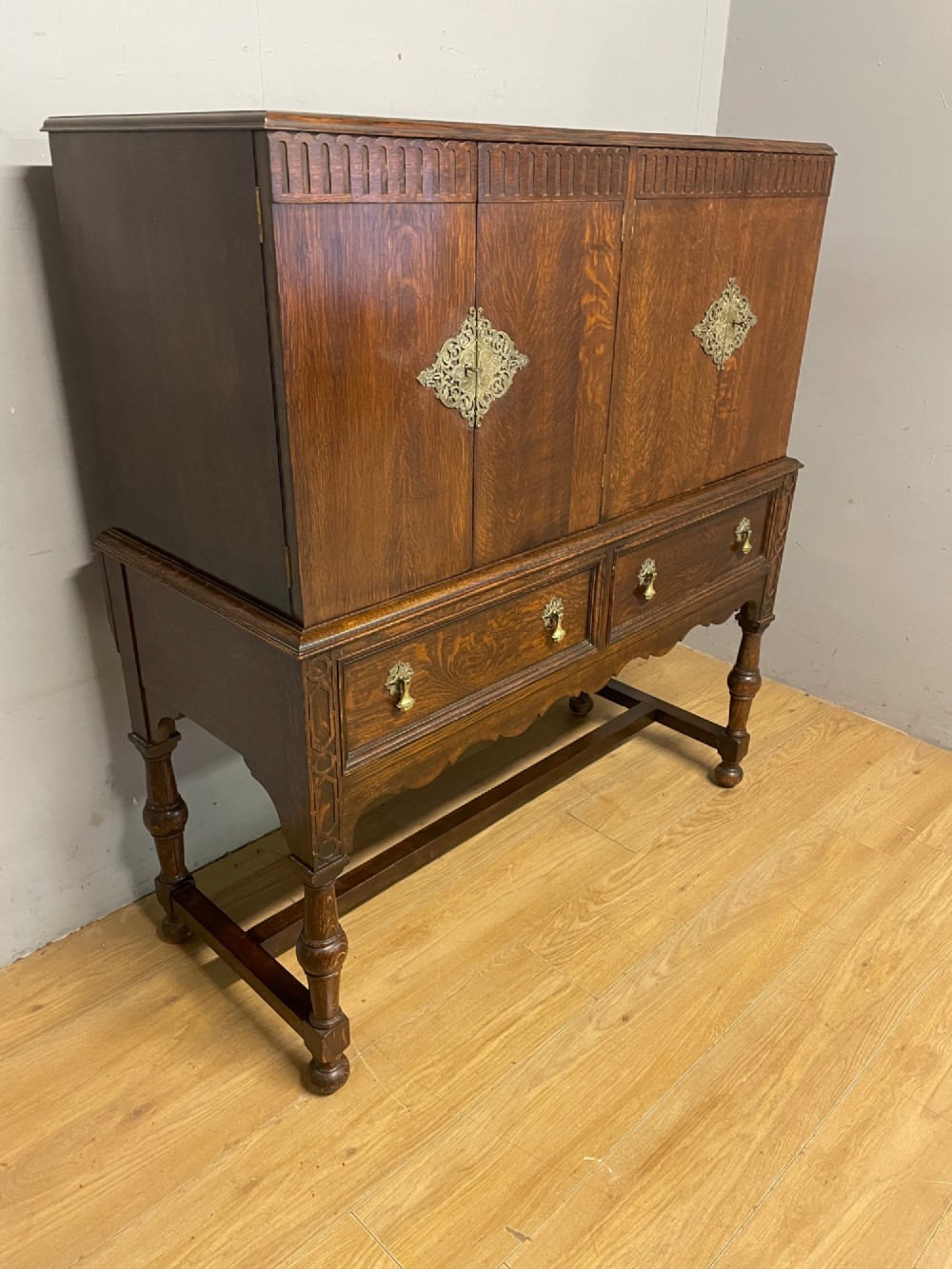 A solid dark oak Jacobean style side cabinet featuring brass escutcheons, carved moulding along the top edge, two lower drawers, and turned legs connected by a lower stretcher. View III