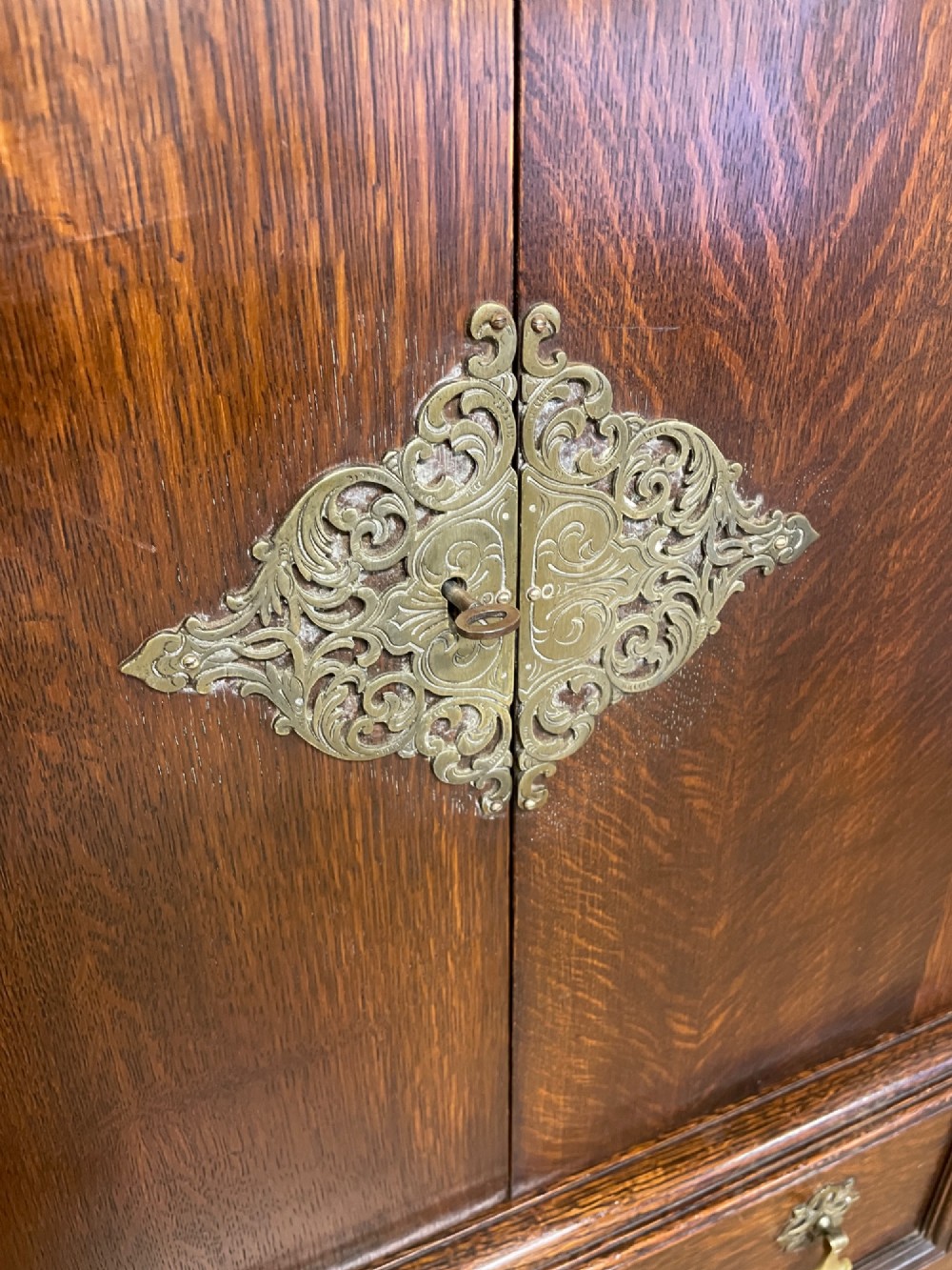 A solid dark oak Jacobean style side cabinet featuring brass escutcheons, carved moulding along the top edge, two lower drawers, and turned legs connected by a lower stretcher. View IV