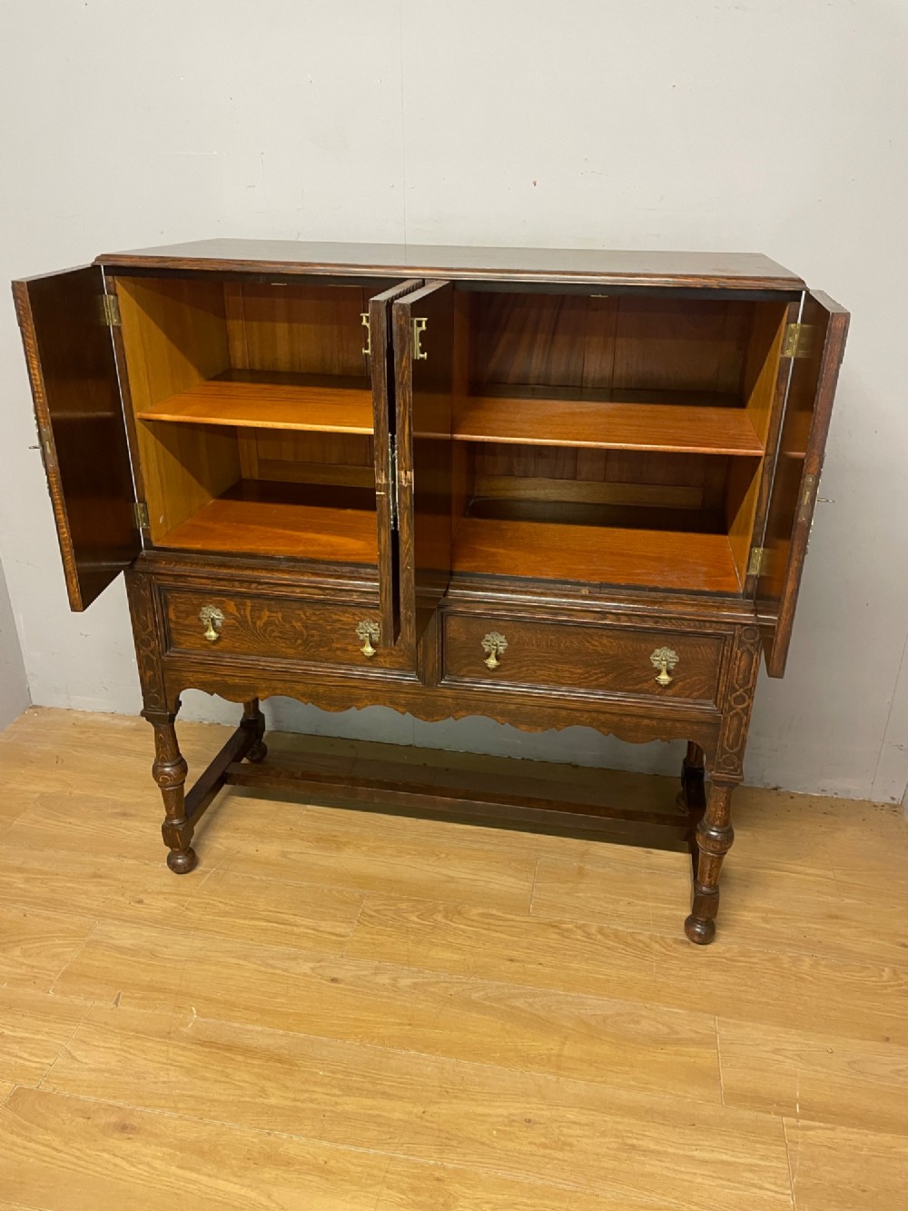 A solid dark oak Jacobean style side cabinet featuring brass escutcheons, carved moulding along the top edge, two lower drawers, and turned legs connected by a lower stretcher. View VI