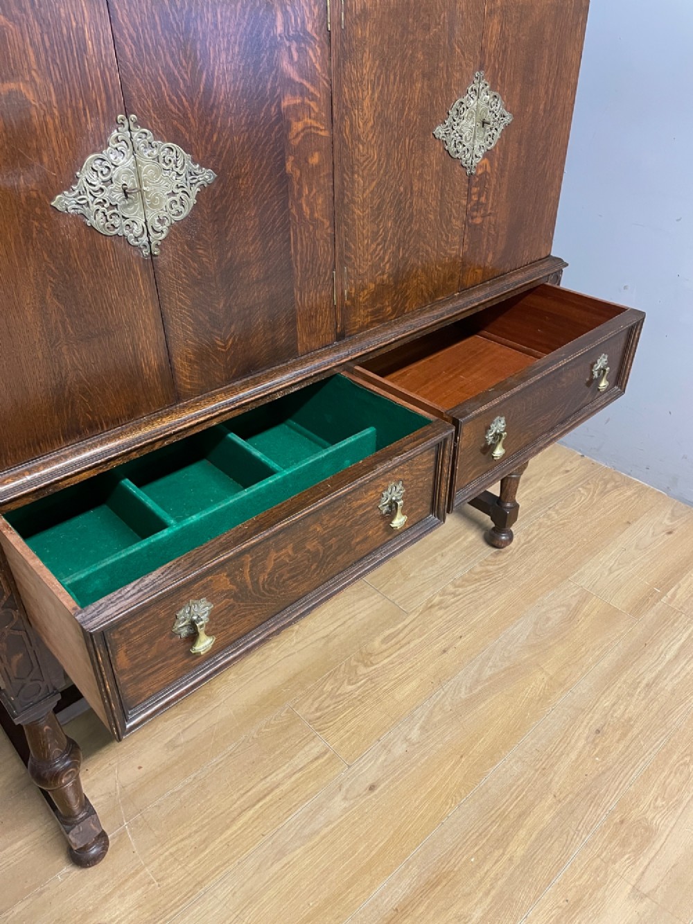 A solid dark oak Jacobean style side cabinet featuring brass escutcheons, carved moulding along the top edge, two lower drawers, and turned legs connected by a lower stretcher. View VII