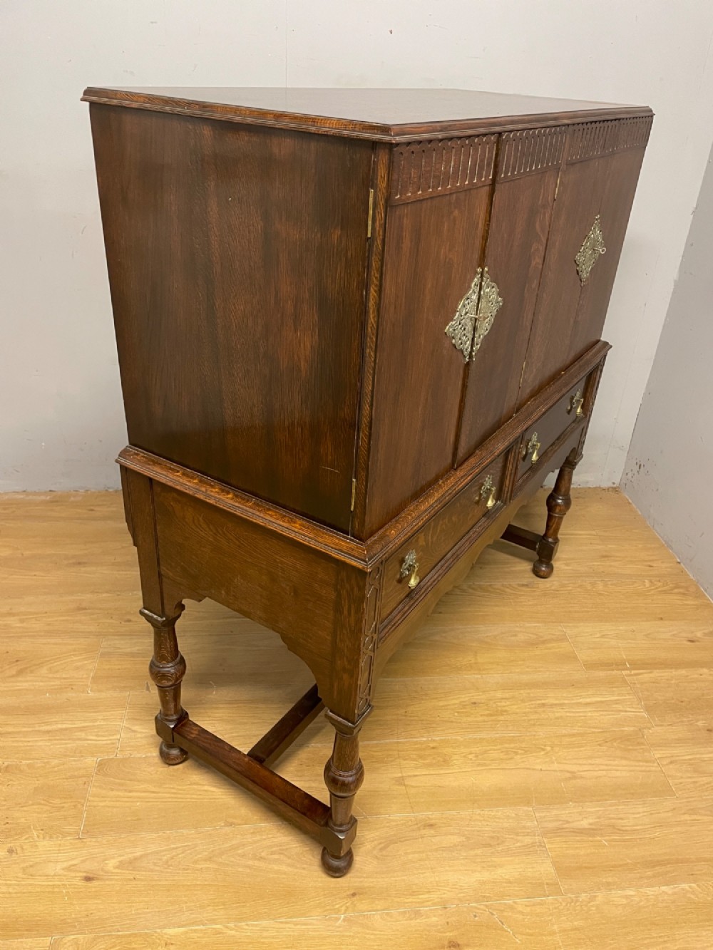 A solid dark oak Jacobean style side cabinet featuring brass escutcheons, carved moulding along the top edge, two lower drawers, and turned legs connected by a lower stretcher. View IX