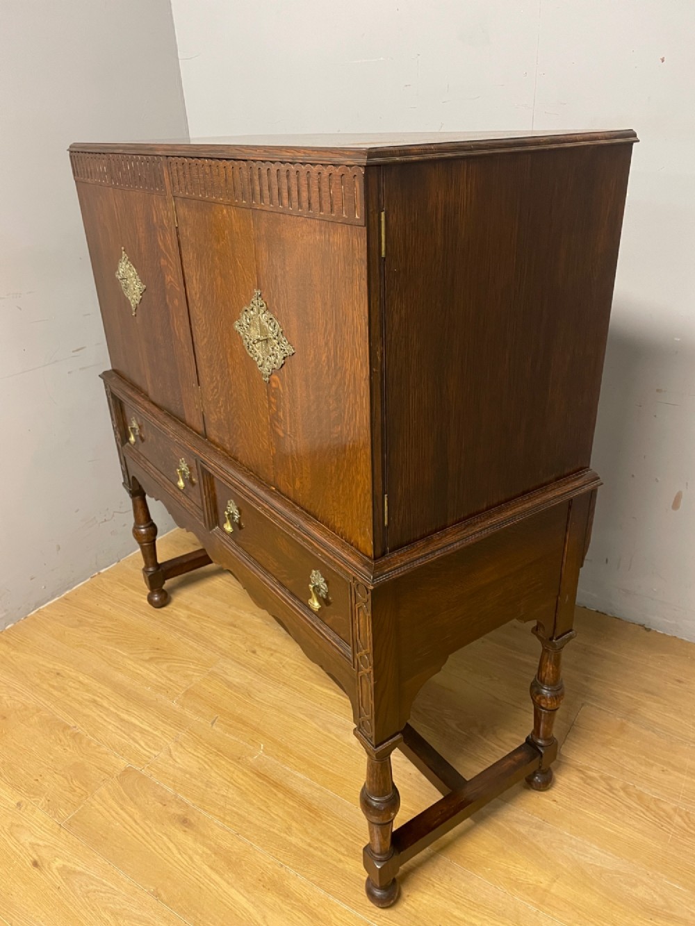A solid dark oak Jacobean style side cabinet featuring brass escutcheons, carved moulding along the top edge, two lower drawers, and turned legs connected by a lower stretcher. View X