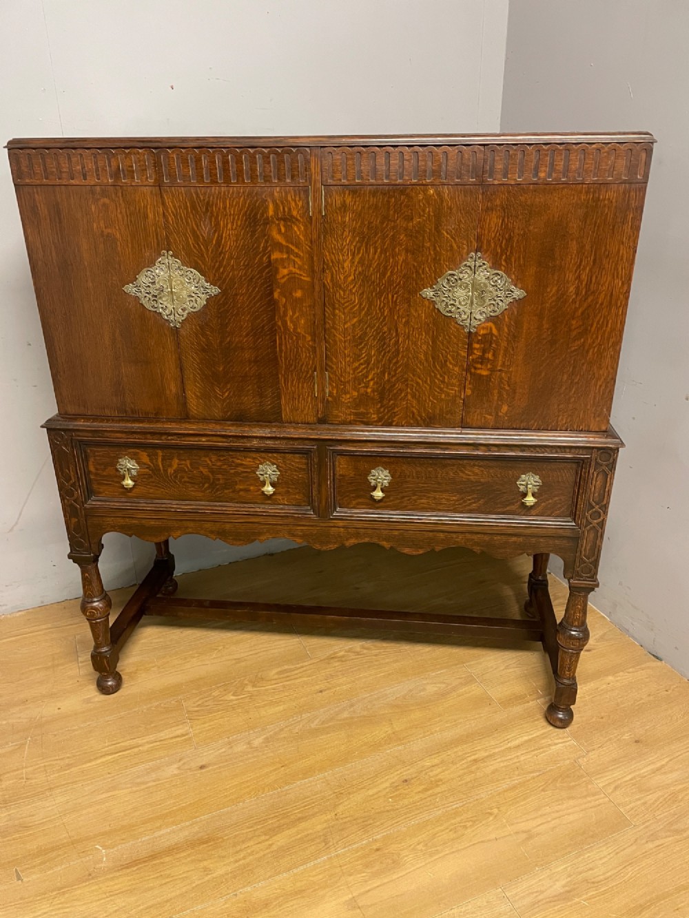 A solid dark oak Jacobean style side cabinet featuring brass escutcheons, carved moulding along the top edge, two lower drawers, and turned legs connected by a lower stretcher. View XII