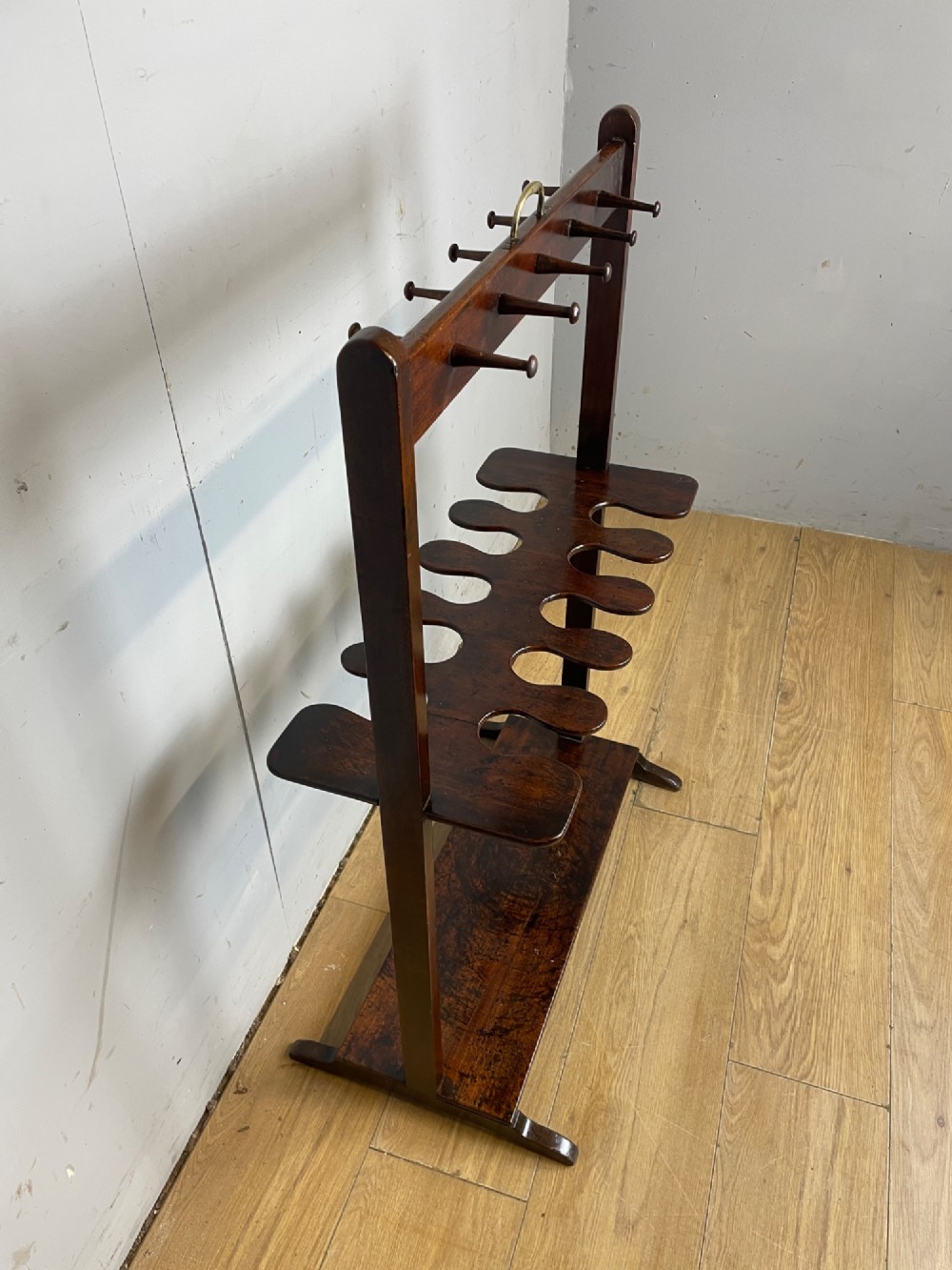 A dark mahogany Georgian boot and whip rack featuring an upper row of turned pegs, a brass handle, a central cutout shelf, and a solid base. View X