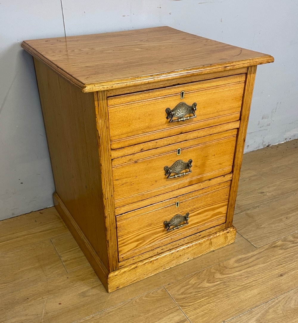 A late 19th-century small ash three-drawer chest featuring cast metal handles, brass keyhole escutcheons, a moulded edge top, and a solid plinth base.