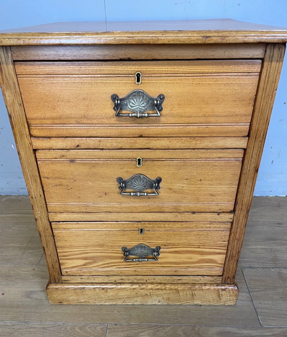 A late 19th-century small ash three-drawer chest featuring cast metal handles, brass keyhole escutcheons, a moulded edge top, and a solid plinth base. View IV