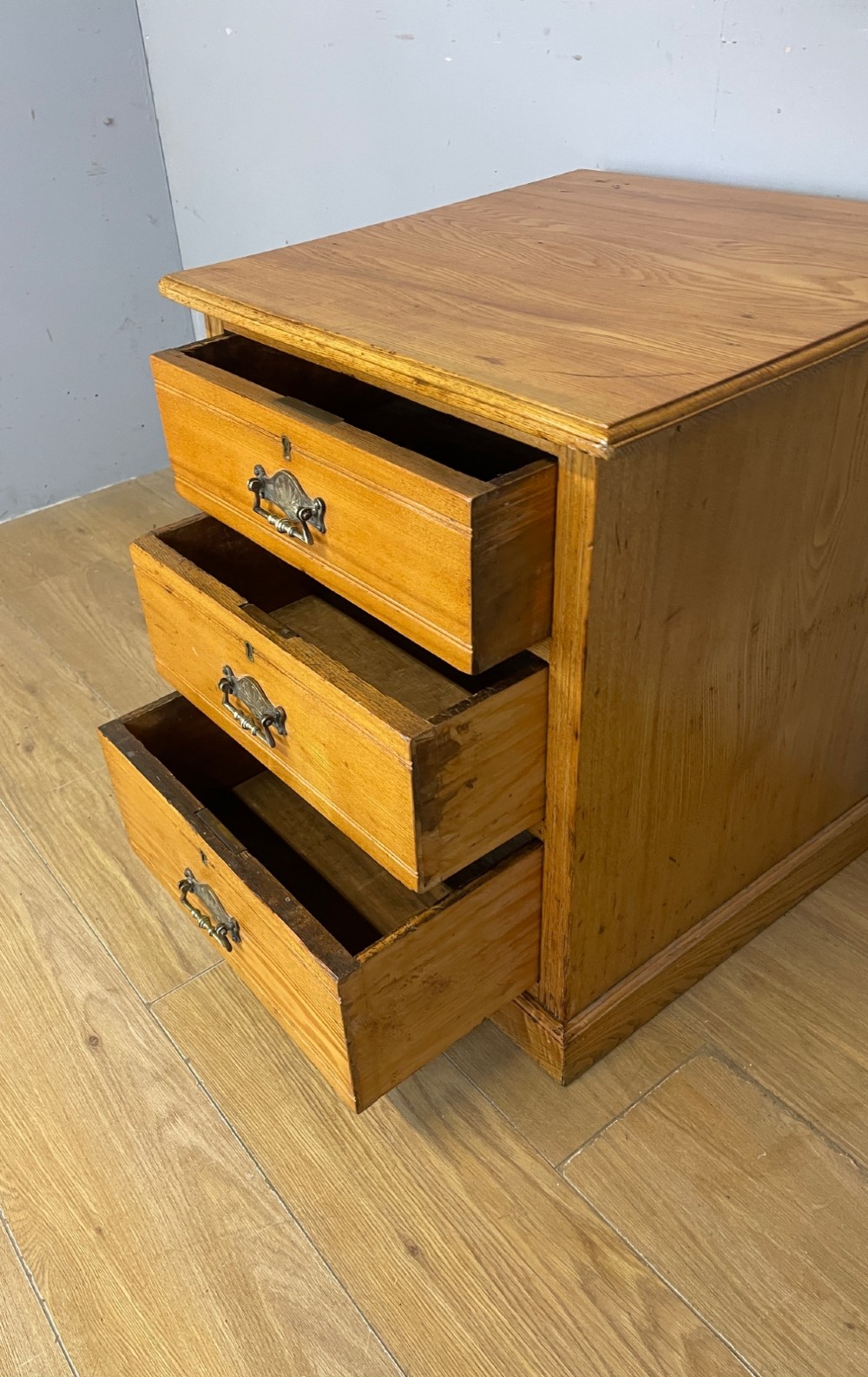 A late 19th-century small ash three-drawer chest featuring cast metal handles, brass keyhole escutcheons, a moulded edge top, and a solid plinth base. View V