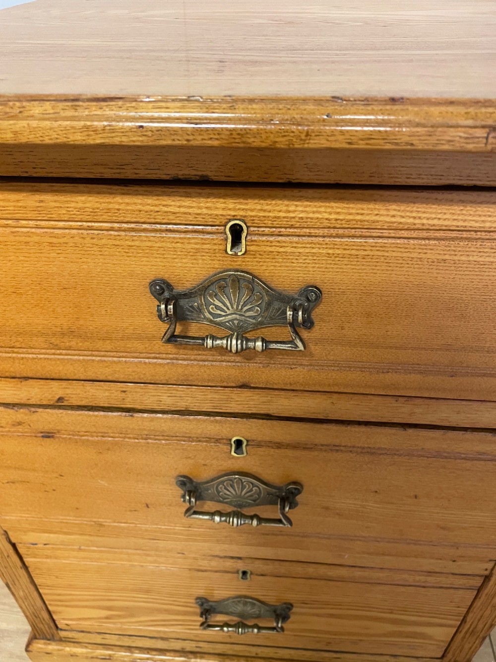 A late 19th-century small ash three-drawer chest featuring cast metal handles, brass keyhole escutcheons, a moulded edge top, and a solid plinth base. View VI