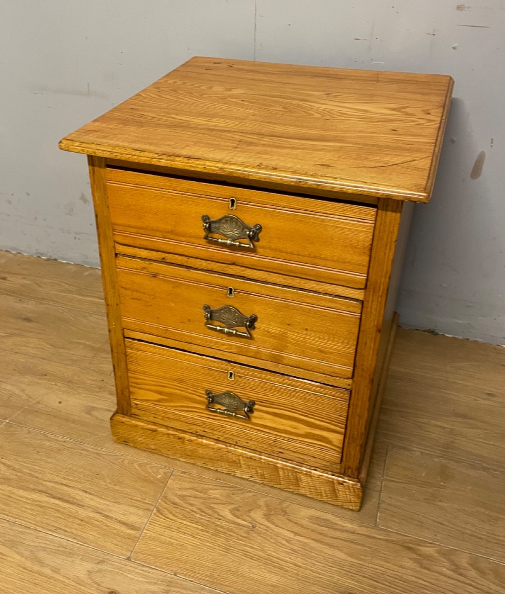 A late 19th-century small ash three-drawer chest featuring cast metal handles, brass keyhole escutcheons, a moulded edge top, and a solid plinth base. View X