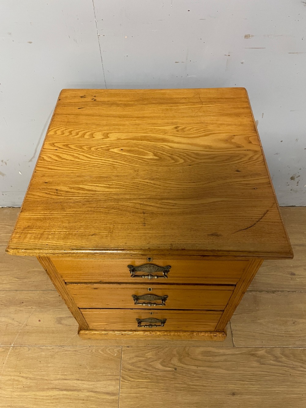 A late 19th-century small ash three-drawer chest featuring cast metal handles, brass keyhole escutcheons, a moulded edge top, and a solid plinth base. View XI