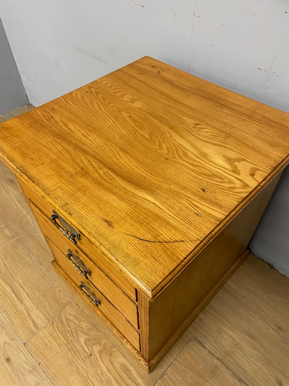 A late 19th-century small ash three-drawer chest featuring cast metal handles, brass keyhole escutcheons, a moulded edge top, and a solid plinth base. View XII