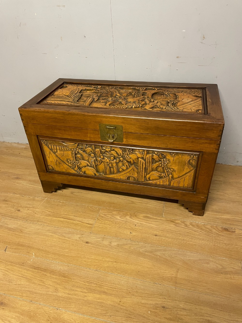 A warm brown rectangular camphor wood chest featuring relief-carved panels depicting figures and landscapes, completed with a central brass latch.