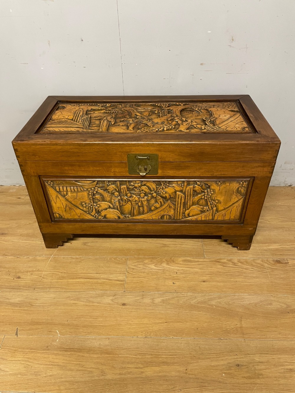 A warm brown rectangular camphor wood chest featuring relief-carved panels depicting figures and landscapes, completed with a central brass latch. View II