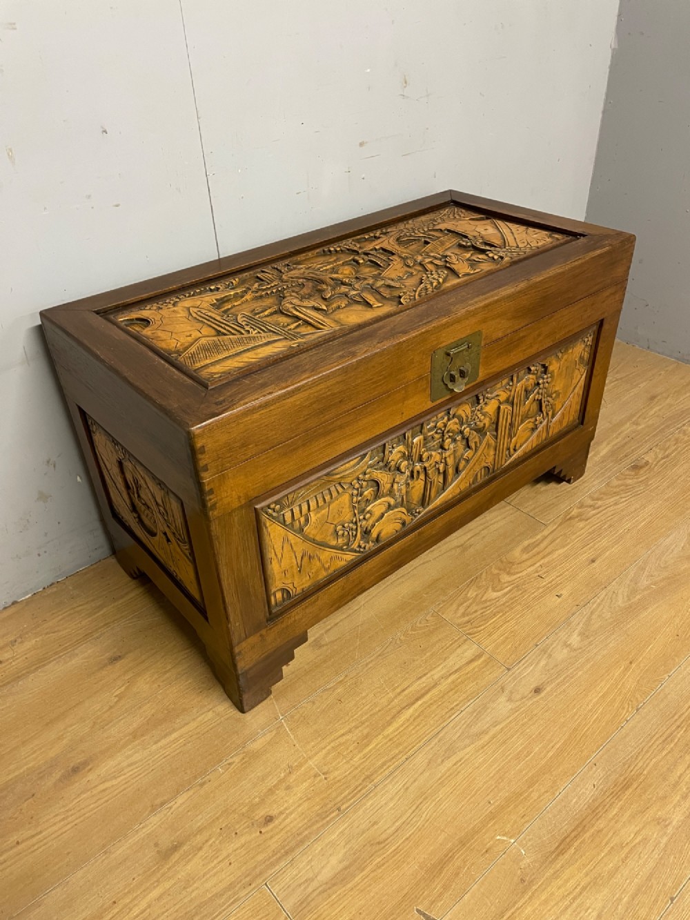 A warm brown rectangular camphor wood chest featuring relief-carved panels depicting figures and landscapes, completed with a central brass latch. View III