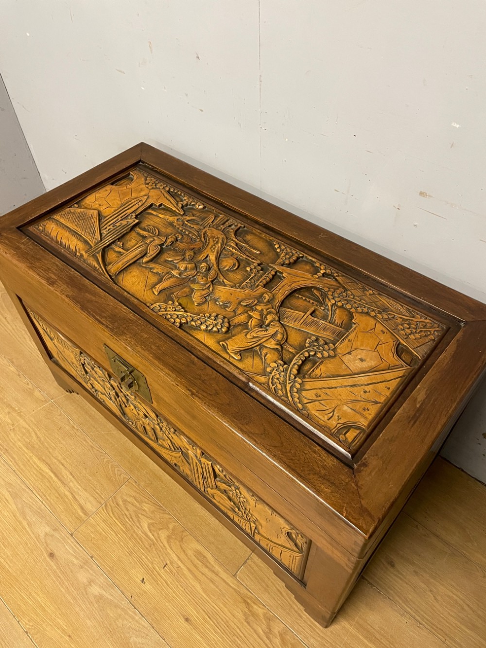 A warm brown rectangular camphor wood chest featuring relief-carved panels depicting figures and landscapes, completed with a central brass latch. View IV