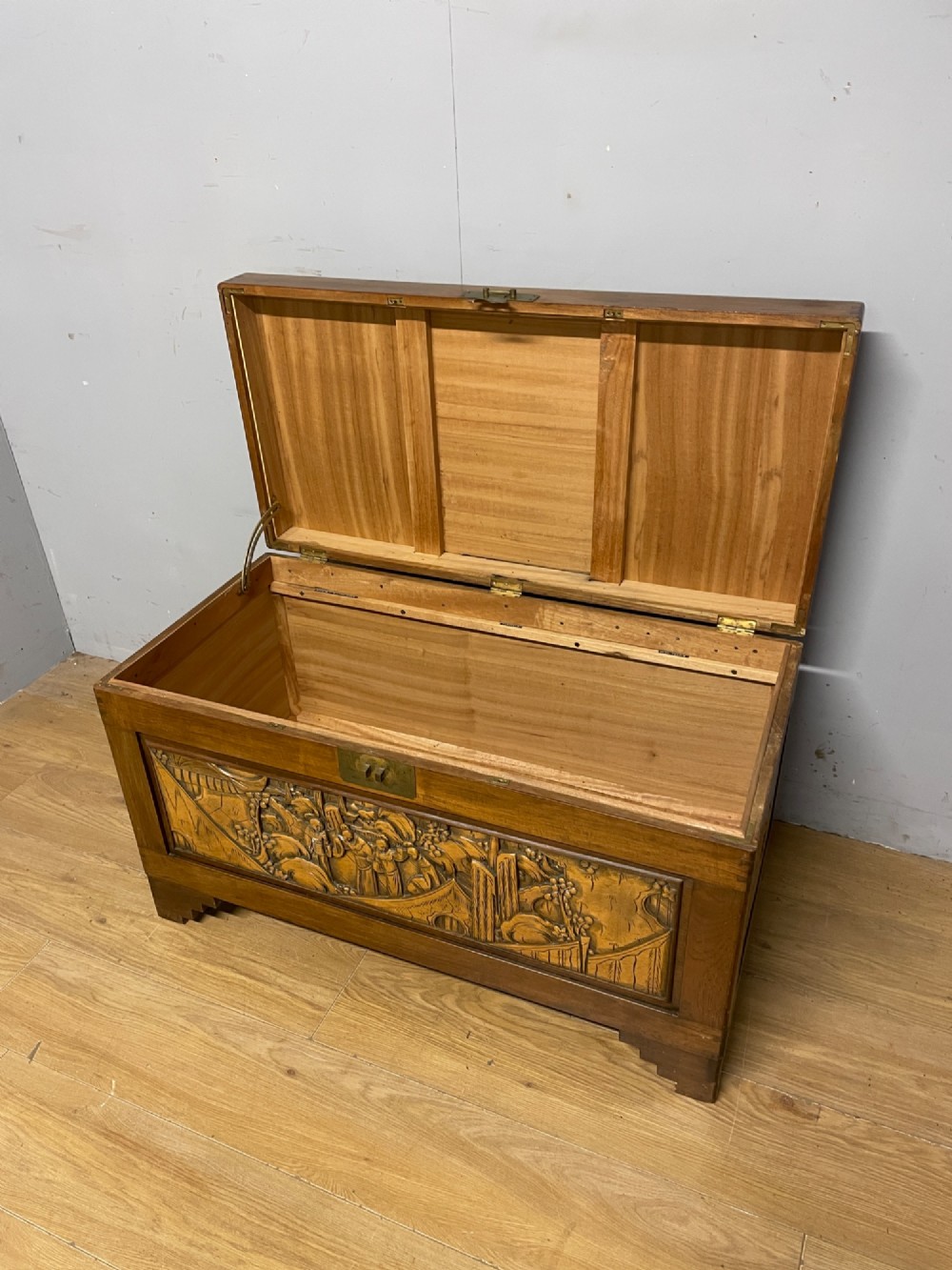 A warm brown rectangular camphor wood chest featuring relief-carved panels depicting figures and landscapes, completed with a central brass latch. View V