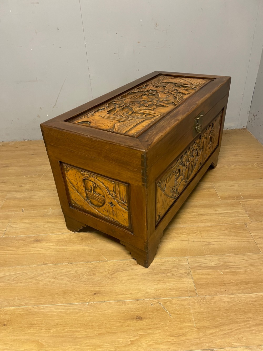 A warm brown rectangular camphor wood chest featuring relief-carved panels depicting figures and landscapes, completed with a central brass latch. View VII
