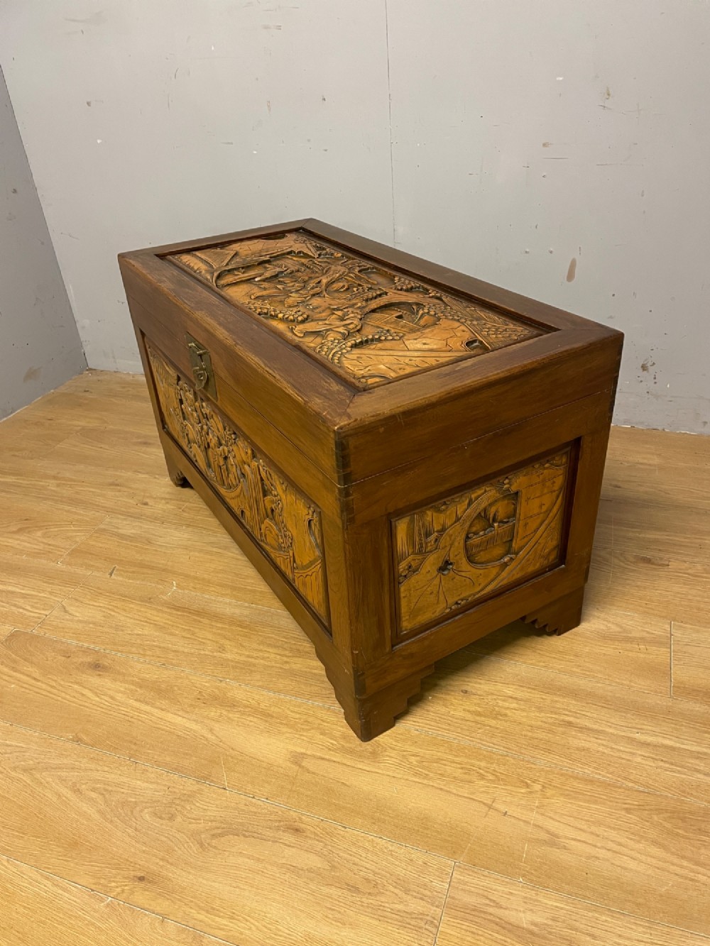 A warm brown rectangular camphor wood chest featuring relief-carved panels depicting figures and landscapes, completed with a central brass latch. View VIII