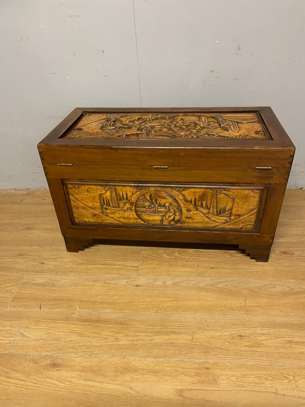 A warm brown rectangular camphor wood chest featuring relief-carved panels depicting figures and landscapes, completed with a central brass latch. View IX