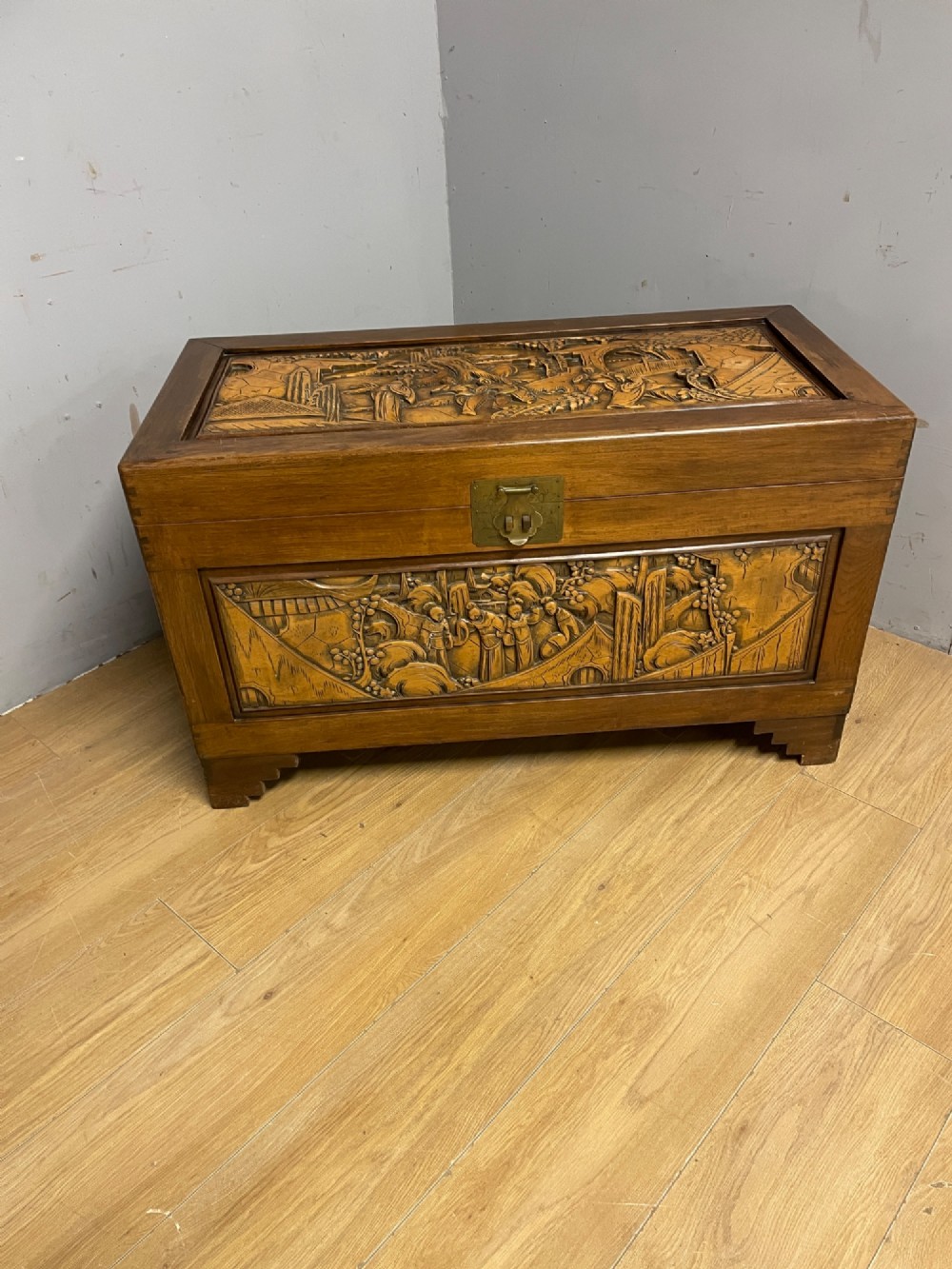 A warm brown rectangular camphor wood chest featuring relief-carved panels depicting figures and landscapes, completed with a central brass latch. View X
