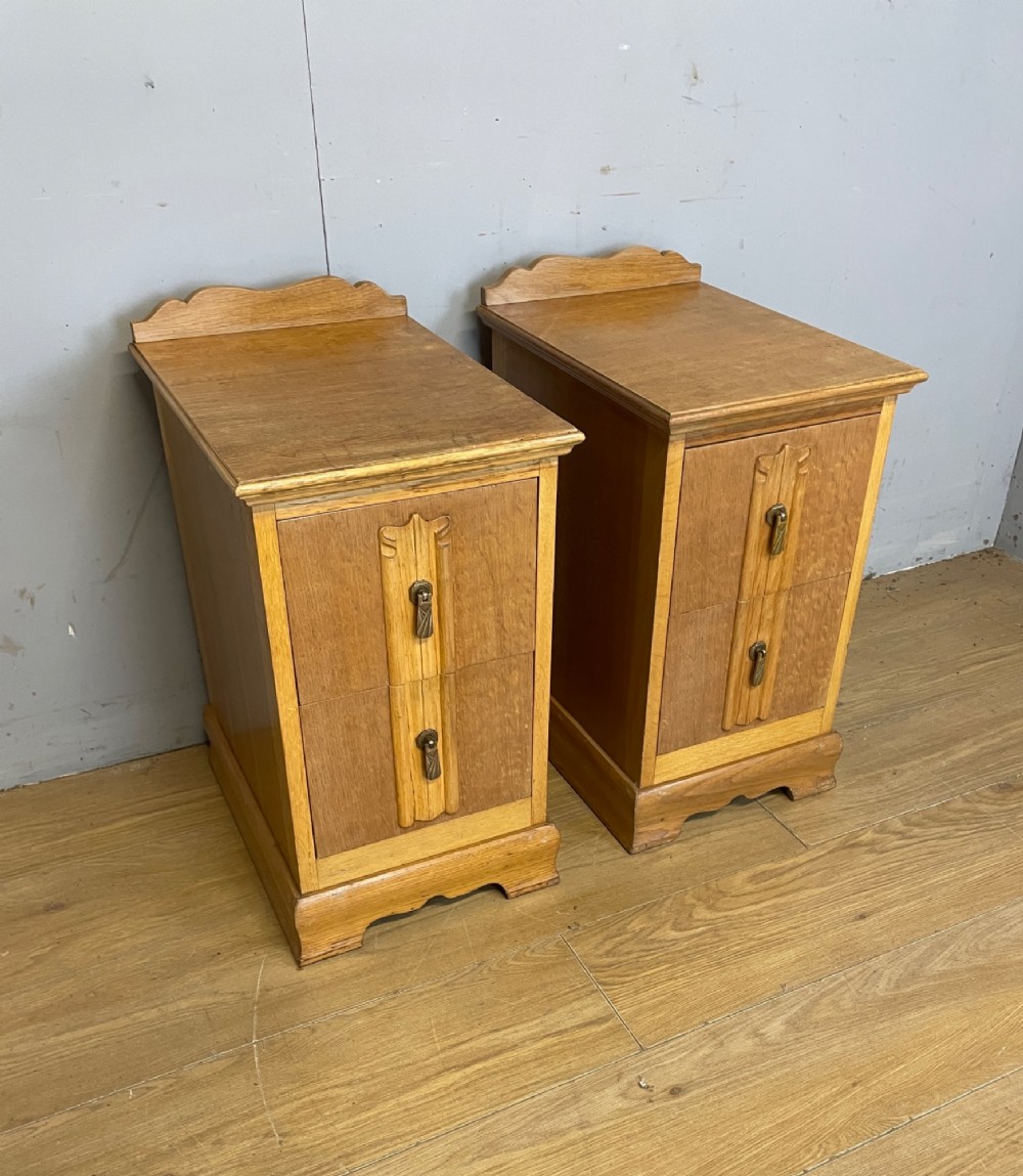 A pair of golden oak Art Deco bedside cabinets featuring two drawers each with stylized carved accents, drop handles, and a low curved back gallery.