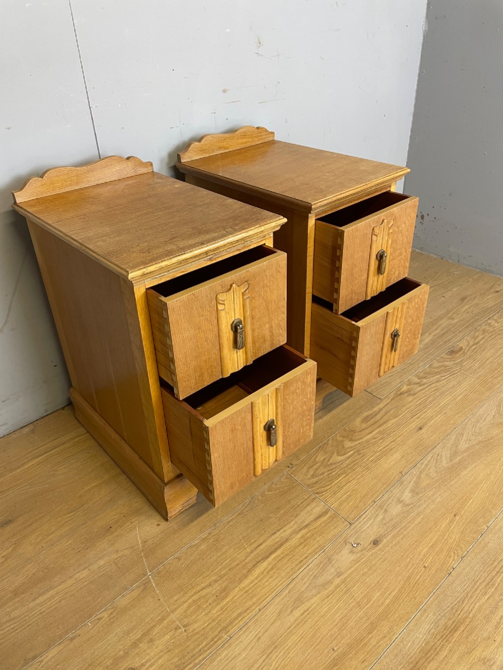 A pair of golden oak Art Deco bedside cabinets featuring two drawers each with stylized carved accents, drop handles, and a low curved back gallery. View VI