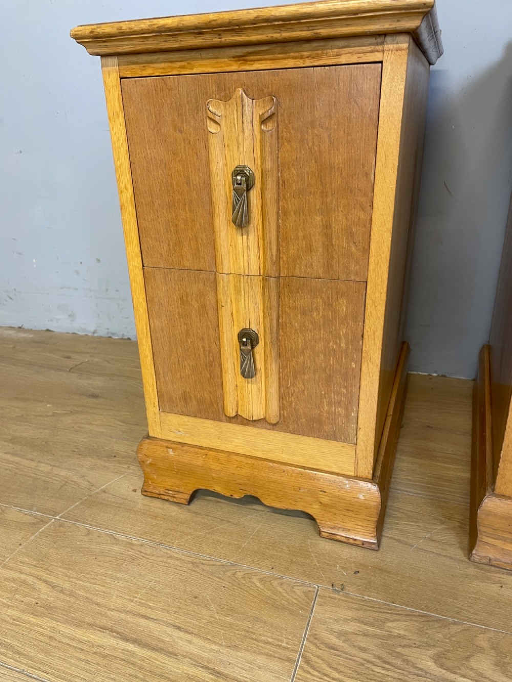 A pair of golden oak Art Deco bedside cabinets featuring two drawers each with stylized carved accents, drop handles, and a low curved back gallery. View X