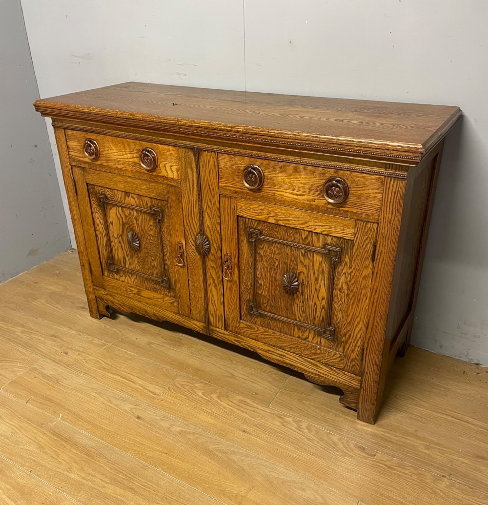 A golden oak two-door sideboard featuring copper drop handles, upper drawers, and carved geometric panelled cupboard doors. View II