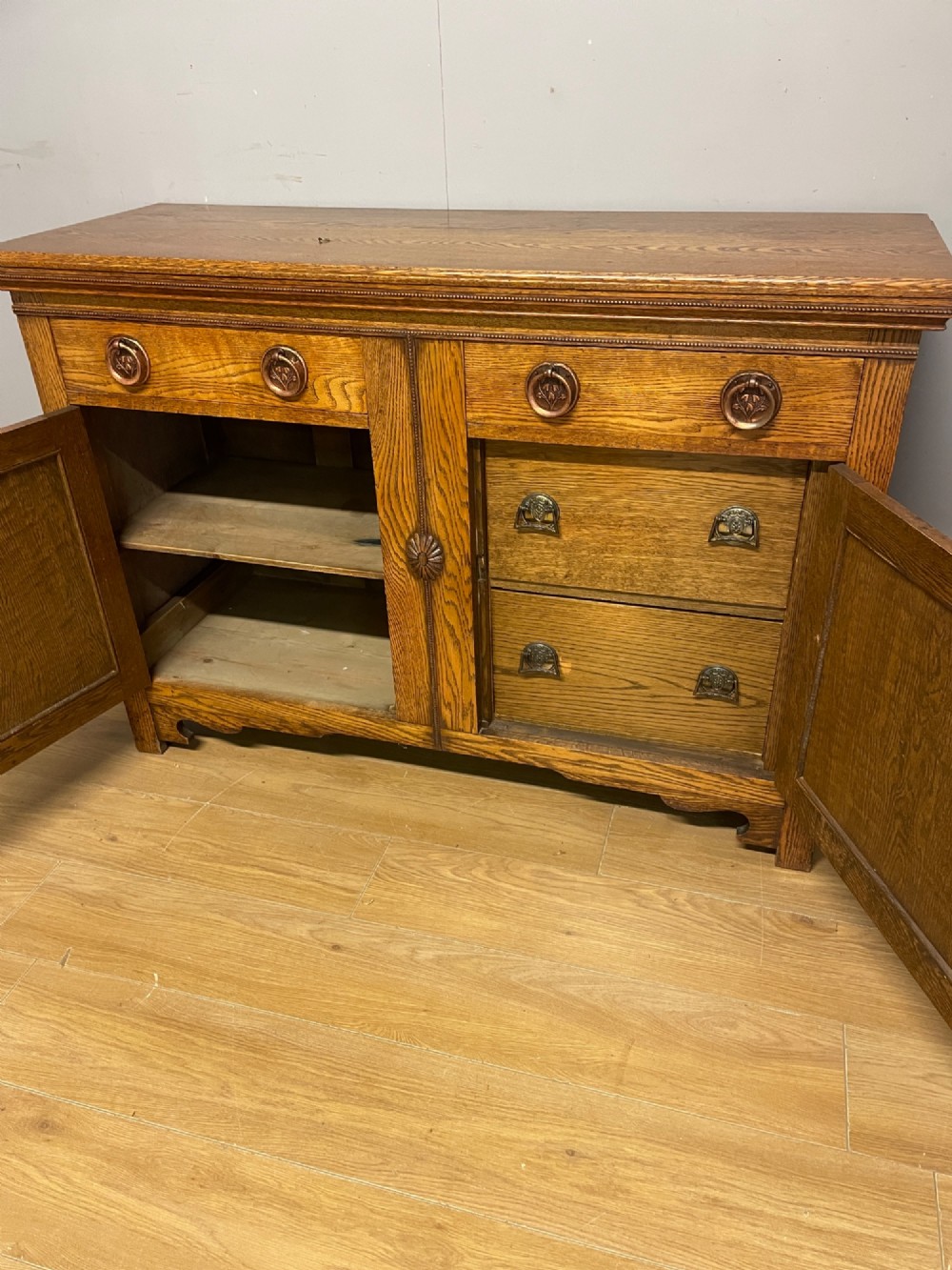A golden oak two-door sideboard featuring copper drop handles, upper drawers, and carved geometric panelled cupboard doors. View V