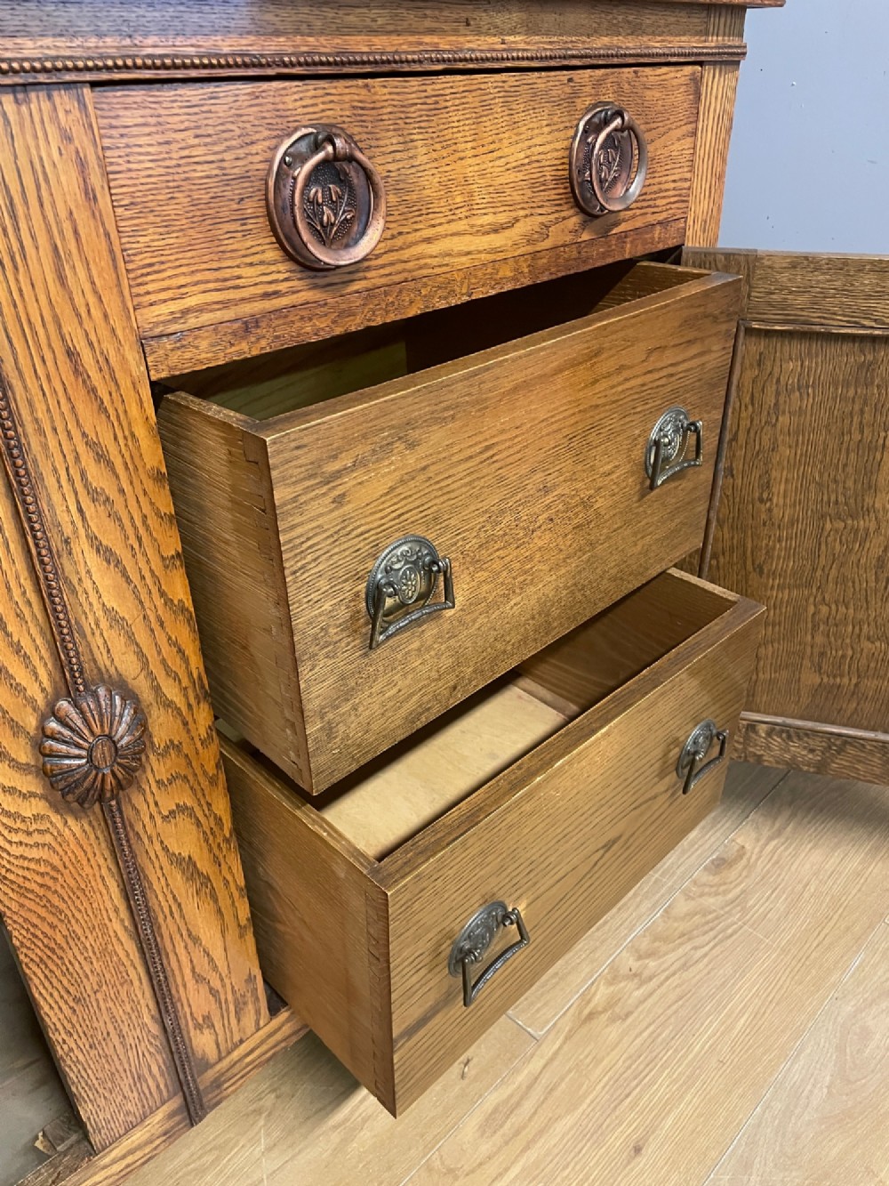 A golden oak two-door sideboard featuring copper drop handles, upper drawers, and carved geometric panelled cupboard doors. View VI