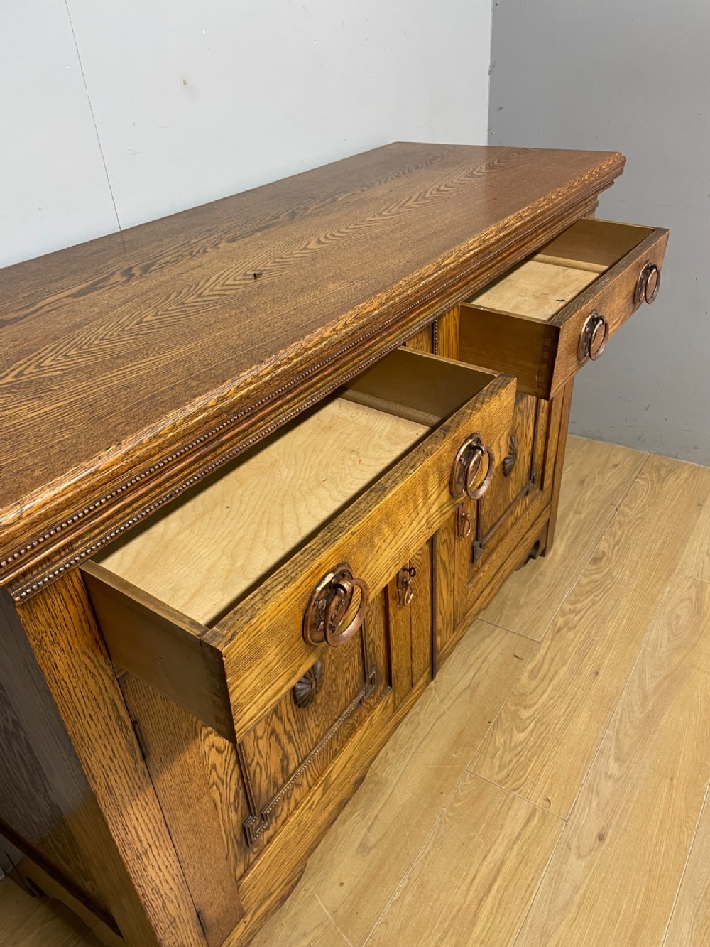 A golden oak two-door sideboard featuring copper drop handles, upper drawers, and carved geometric panelled cupboard doors. View VII