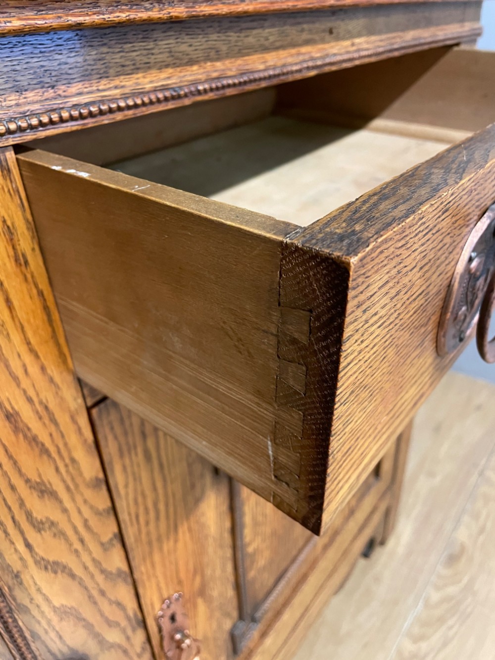 A golden oak two-door sideboard featuring copper drop handles, upper drawers, and carved geometric panelled cupboard doors. View VIII