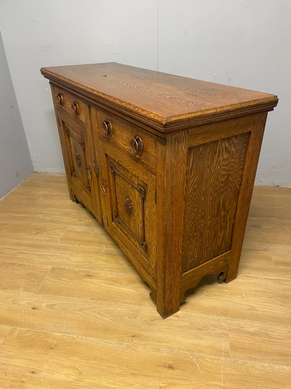 A golden oak two-door sideboard featuring copper drop handles, upper drawers, and carved geometric panelled cupboard doors. View IX