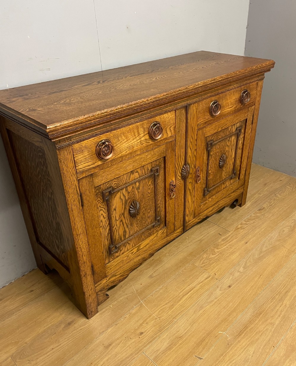 A golden oak two-door sideboard featuring copper drop handles, upper drawers, and carved geometric panelled cupboard doors. View X