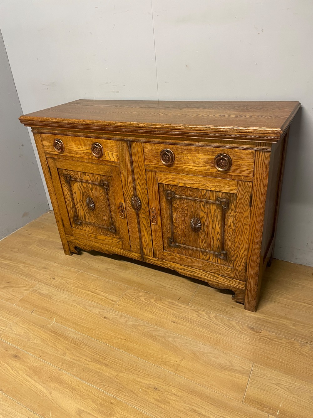 A golden oak two-door sideboard featuring copper drop handles, upper drawers, and carved geometric panelled cupboard doors. View XII