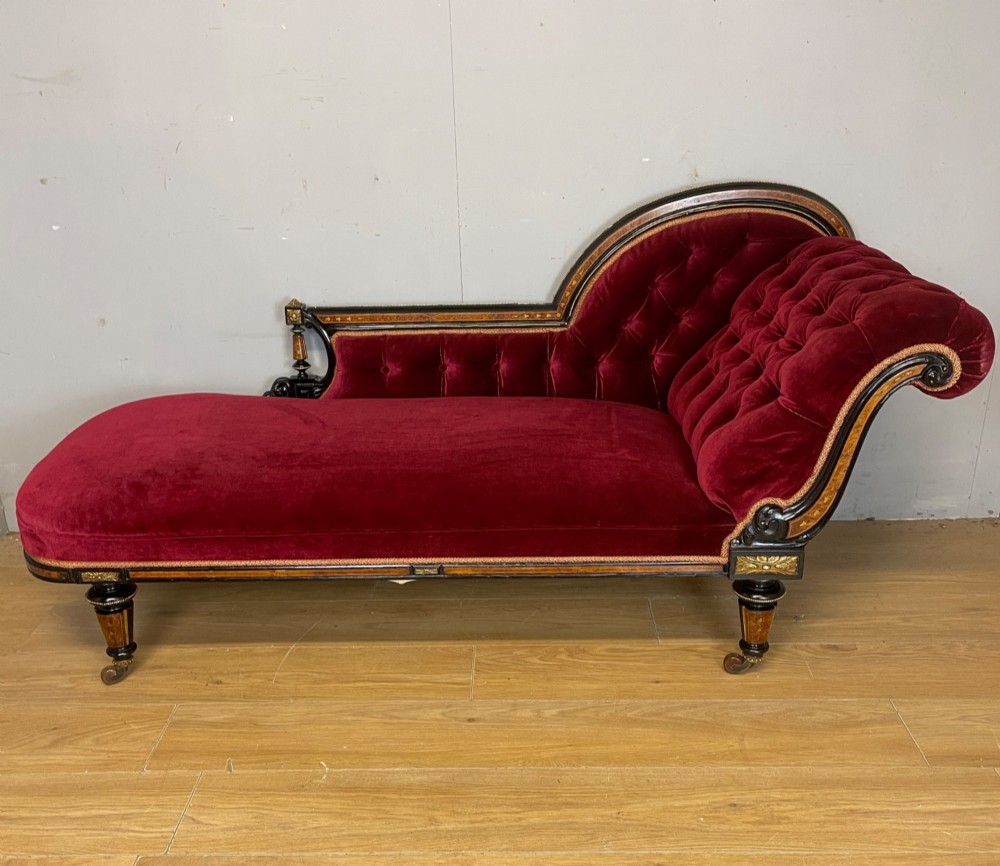 A 19th-century ebonised chaise longue with deep red velvet tufted upholstery, walnut inlaid borders, turned legs on brass castors, and decorative ormolu mounts.