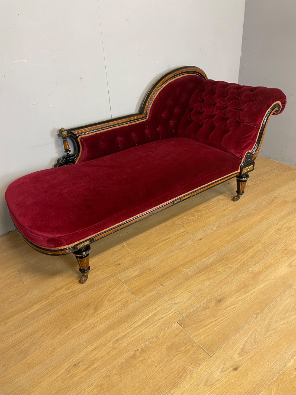 A 19th-century ebonised chaise longue with deep red velvet tufted upholstery, walnut inlaid borders, turned legs on brass castors, and decorative ormolu mounts. View II