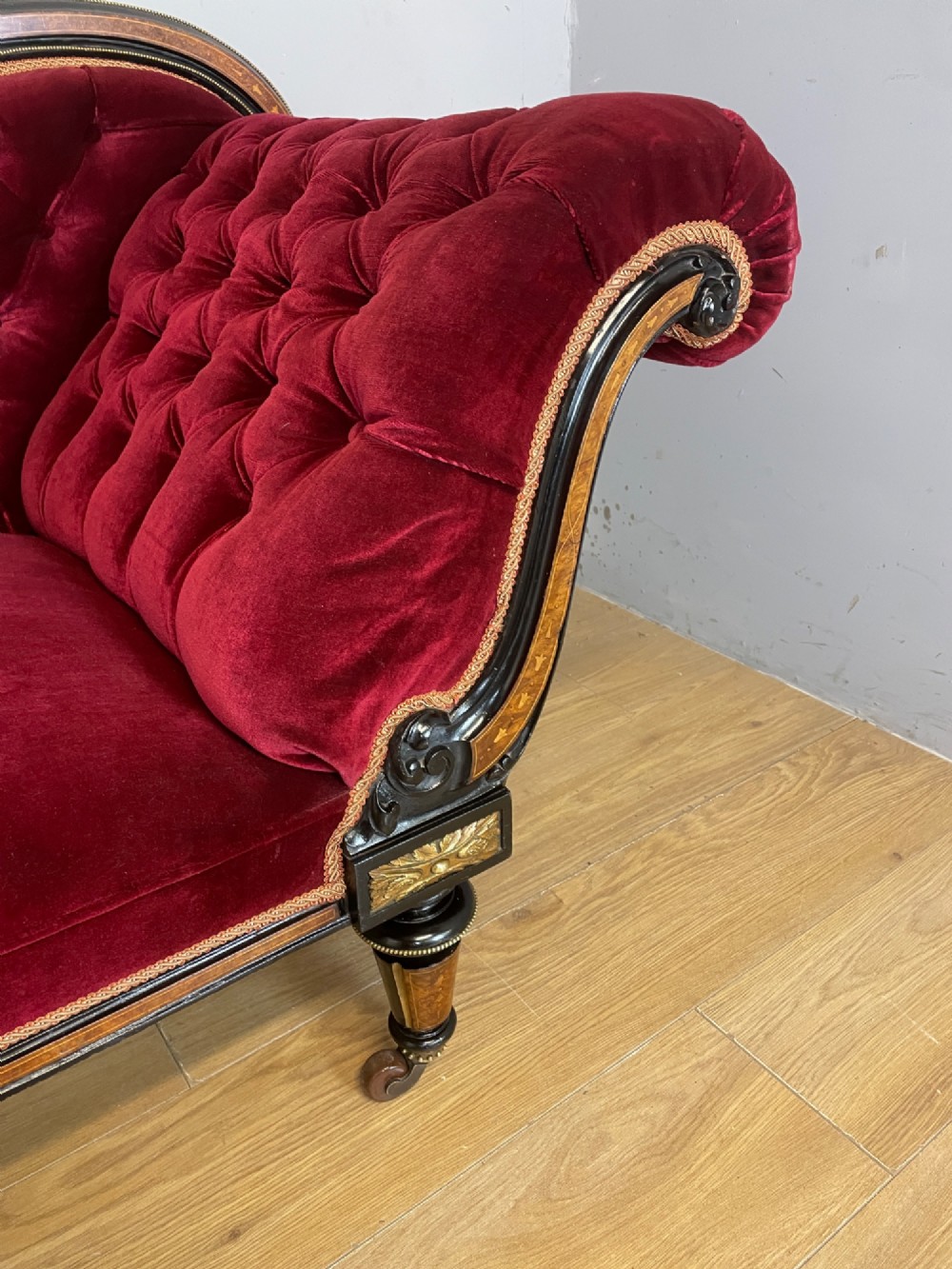 A 19th-century ebonised chaise longue with deep red velvet tufted upholstery, walnut inlaid borders, turned legs on brass castors, and decorative ormolu mounts. View III
