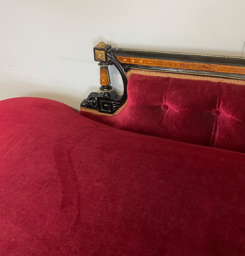 A 19th-century ebonised chaise longue with deep red velvet tufted upholstery, walnut inlaid borders, turned legs on brass castors, and decorative ormolu mounts. View V