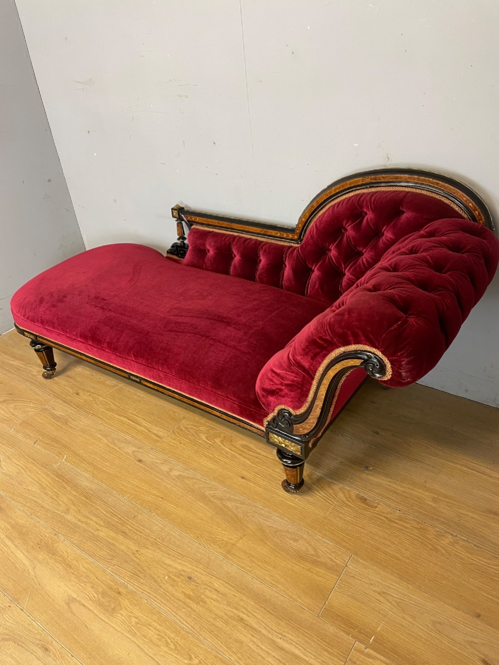 A 19th-century ebonised chaise longue with deep red velvet tufted upholstery, walnut inlaid borders, turned legs on brass castors, and decorative ormolu mounts. View VII