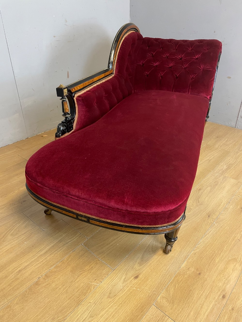 A 19th-century ebonised chaise longue with deep red velvet tufted upholstery, walnut inlaid borders, turned legs on brass castors, and decorative ormolu mounts. View IX