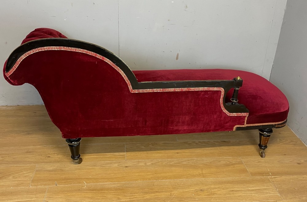 A 19th-century ebonised chaise longue with deep red velvet tufted upholstery, walnut inlaid borders, turned legs on brass castors, and decorative ormolu mounts. View X